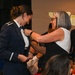 A formal military commissioning or promotion ceremony is taking place in a banquet hall setting. A woman in civilian attire is pinning insignia or a device onto the collar of a person wearing a dark blue military dress uniform. Both individuals are smiling, suggesting a celebratory and emotionally significant moment.
