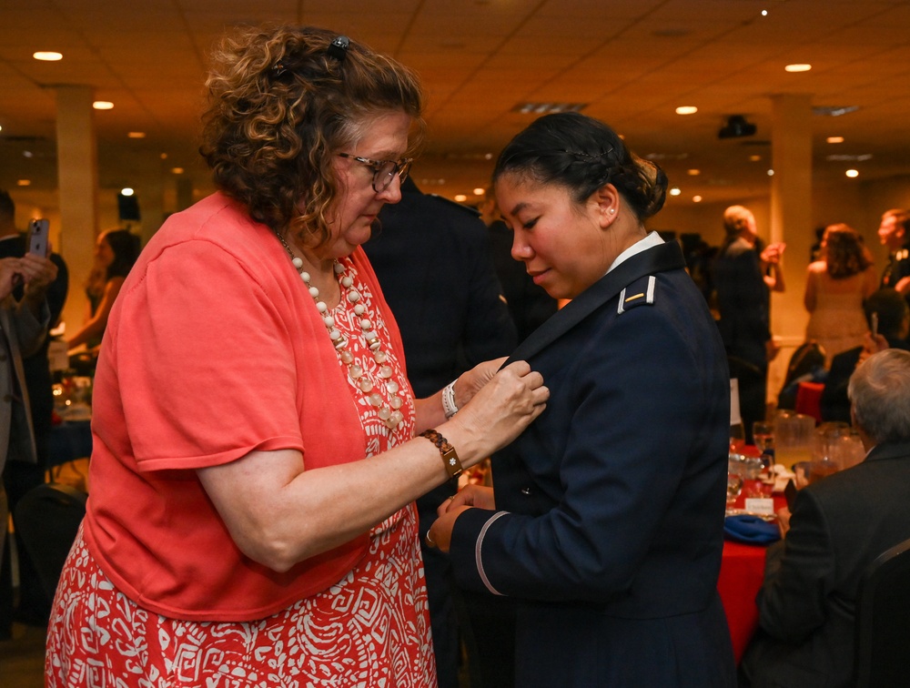 A woman in civilian clothing is pinning something onto the uniform of a military officer during what appears to be a formal commissioning or promotion ceremony. The officer is wearing a dark blue military dress uniform with a single gold bar insignia visible on the shoulder, suggesting the rank of Second Lieutenant or Ensign. The event is taking place in a banquet hall with other attendees visible in the background.