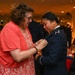 A woman in civilian clothing is pinning something onto the uniform of a military officer during what appears to be a formal commissioning or promotion ceremony. The officer is wearing a dark blue military dress uniform with a single gold bar insignia visible on the shoulder, suggesting the rank of Second Lieutenant or Ensign. The event is taking place in a banquet hall with other attendees visible in the background.