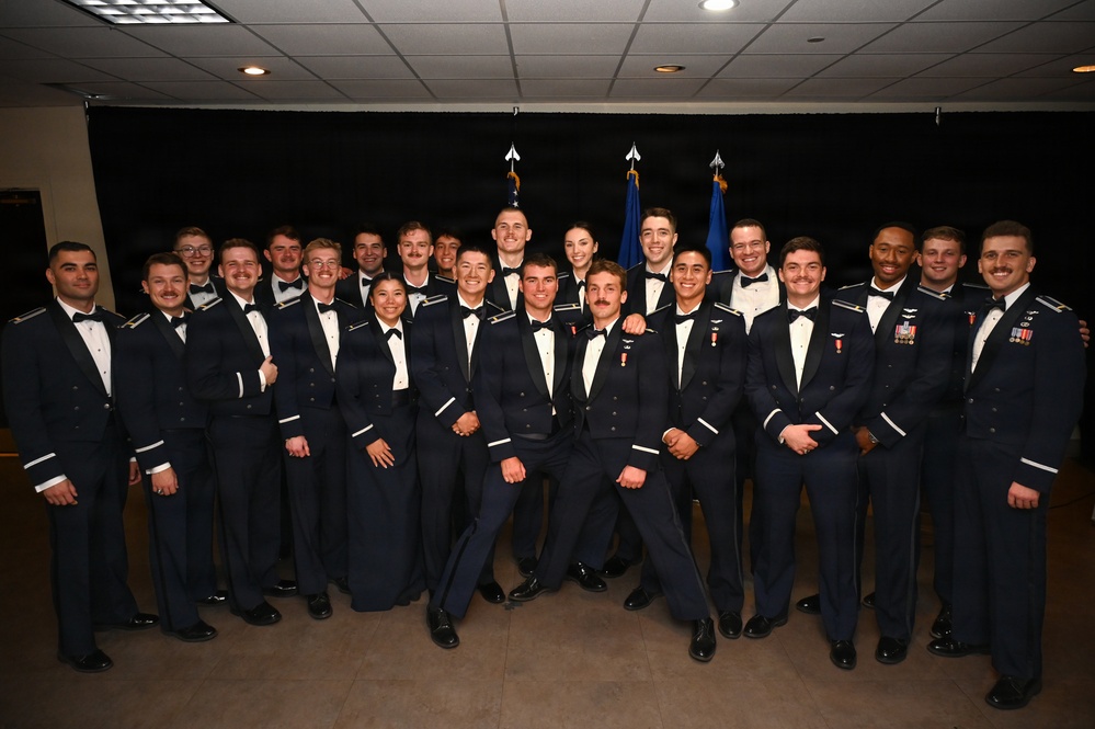 A large group of approximately 25 military personnel posed for a formal group photograph in dress uniforms. The individuals are wearing U.S. Air Force mess dress uniforms (dark navy blue with white dress shirts and bow ties), suggesting a formal military ceremony or graduation event. Two flags are visible in the background, consistent with a U.S. Air Force formal occasion.