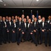 A large group of approximately 25 military personnel posed for a formal group photograph in dress uniforms. The individuals are wearing U.S. Air Force mess dress uniforms (dark navy blue with white dress shirts and bow ties), suggesting a formal military ceremony or graduation event. Two flags are visible in the background, consistent with a U.S. Air Force formal occasion.