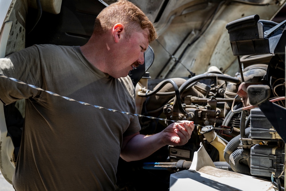 A young man with red hair wearing an olive/military green t-shirt is working on a vehicle engine with the hood open. He appears to be inspecting or manipulating a component, possibly checking a dipstick or handling a connector/wire. The vehicle's engine bay shows significant wear and dust, suggesting a military or heavy-use vehicle.