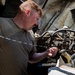 A young man with red hair wearing an olive/military green t-shirt is working on a vehicle engine with the hood open. He appears to be inspecting or manipulating a component, possibly checking a dipstick or handling a connector/wire. The vehicle's engine bay shows significant wear and dust, suggesting a military or heavy-use vehicle.