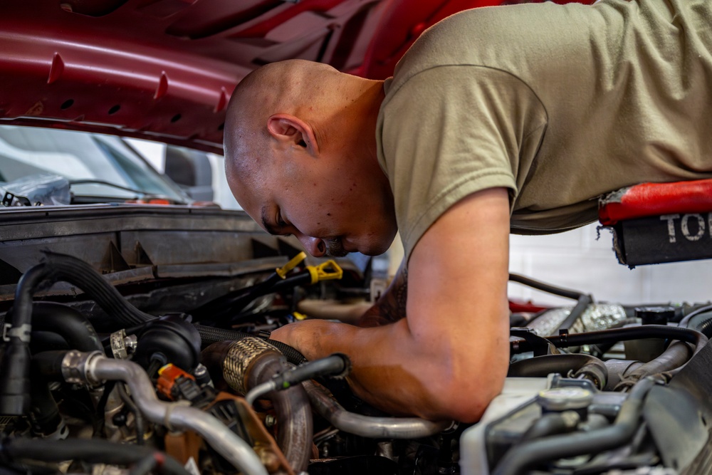 A bald man wearing a tan/olive t-shirt is leaning over and working on a vehicle engine bay. He appears to be inspecting or repairing components within the engine compartment of a red vehicle. The image is taken in what appears to be an automotive workshop or garage setting.