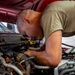 A bald man wearing a tan/olive t-shirt is leaning over and working on a vehicle engine bay. He appears to be inspecting or repairing components within the engine compartment of a red vehicle. The image is taken in what appears to be an automotive workshop or garage setting.
