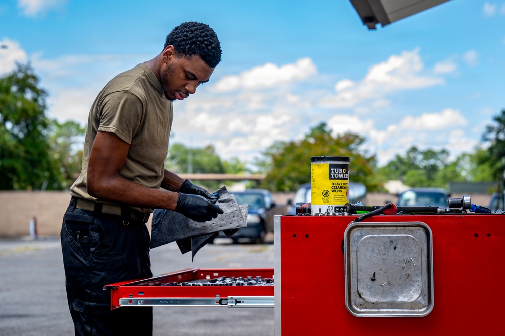 A young man wearing black gloves and an olive green t-shirt is working at a red tool chest outdoors in a parking lot. He appears to be cleaning or handling tools using heavy-duty wipes from a 'Tub O' Towels' container visible on top of the tool chest. The setting suggests a mobile mechanical or maintenance work environment.