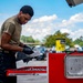 A young man wearing black gloves and an olive green t-shirt is working at a red tool chest outdoors in a parking lot. He appears to be cleaning or handling tools using heavy-duty wipes from a 'Tub O' Towels' container visible on top of the tool chest. The setting suggests a mobile mechanical or maintenance work environment.