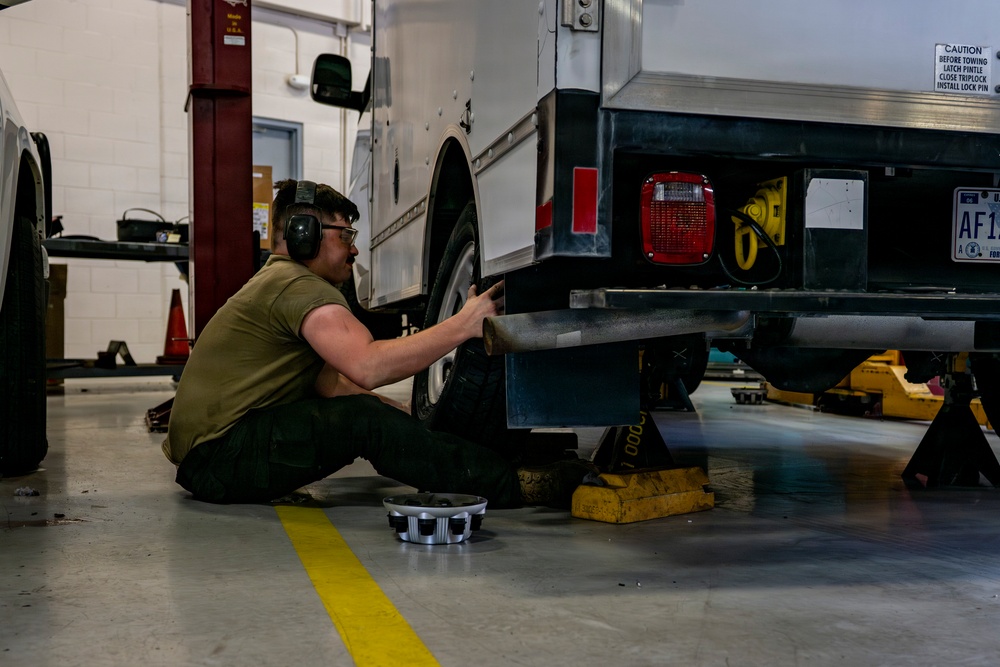 A mechanic is working on the underside of a military or government vehicle in an indoor automotive repair bay. The worker is seated on the floor wearing hearing protection and safety glasses while handling what appears to be an exhaust component. The vehicle is supported by yellow jack stands and shows a license plate beginning with 'AF1', suggesting Air Force affiliation.