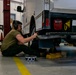 A mechanic is working on the underside of a military or government vehicle in an indoor automotive repair bay. The worker is seated on the floor wearing hearing protection and safety glasses while handling what appears to be an exhaust component. The vehicle is supported by yellow jack stands and shows a license plate beginning with 'AF1', suggesting Air Force affiliation.