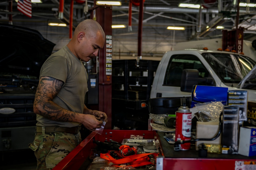 A military service member in olive drab t-shirt and camouflage pants works at a red tool cart in what appears to be a military vehicle maintenance bay. The individual has a shaved head and visible tattoos on their right arm and is handling small parts or components. The background shows a large garage facility with vehicles, fluorescent lighting, and an American flag.