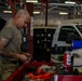 A military service member in olive drab t-shirt and camouflage pants works at a red tool cart in what appears to be a military vehicle maintenance bay. The individual has a shaved head and visible tattoos on their right arm and is handling small parts or components. The background shows a large garage facility with vehicles, fluorescent lighting, and an American flag.