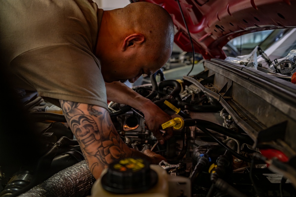 A bald man with extensive tattoos on his right arm is leaning over the open engine bay of a red vehicle, performing mechanical work. He is using a yellow-handled tool to work on engine components in what appears to be an automotive garage or workshop. The image is captured in a dramatic, close-up style with selective lighting emphasizing the subject.