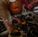 A bald man with extensive tattoos on his right arm is leaning over the open engine bay of a red vehicle, performing mechanical work. He is using a yellow-handled tool to work on engine components in what appears to be an automotive garage or workshop. The image is captured in a dramatic, close-up style with selective lighting emphasizing the subject.