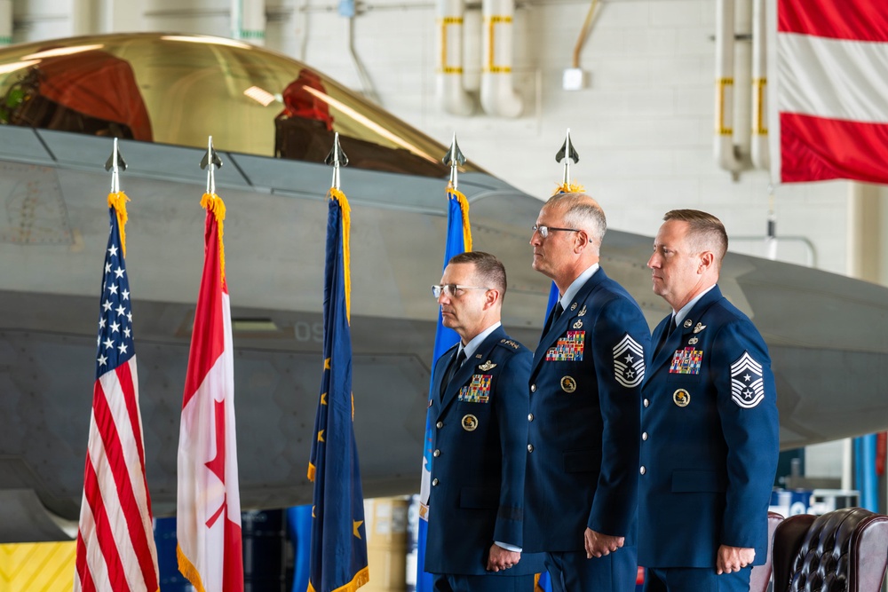 Three U.S. Air Force personnel in dress blue uniforms stand at attention during a formal military ceremony inside an aircraft hangar. A stealth fighter jet (likely an F-22 Raptor) is visible in the background along with multiple flags including the U.S. flag, Canadian flag, and what appears to be a state flag. The two rightmost individuals display Chief Master Sergeant rank insignia, while the leftmost appears to be a commissioned officer.