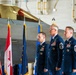 Three U.S. Air Force personnel in dress blue uniforms stand at attention during a formal military ceremony inside an aircraft hangar. A stealth fighter jet (likely an F-22 Raptor) is visible in the background along with multiple flags including the U.S. flag, Canadian flag, and what appears to be a state flag. The two rightmost individuals display Chief Master Sergeant rank insignia, while the leftmost appears to be a commissioned officer.