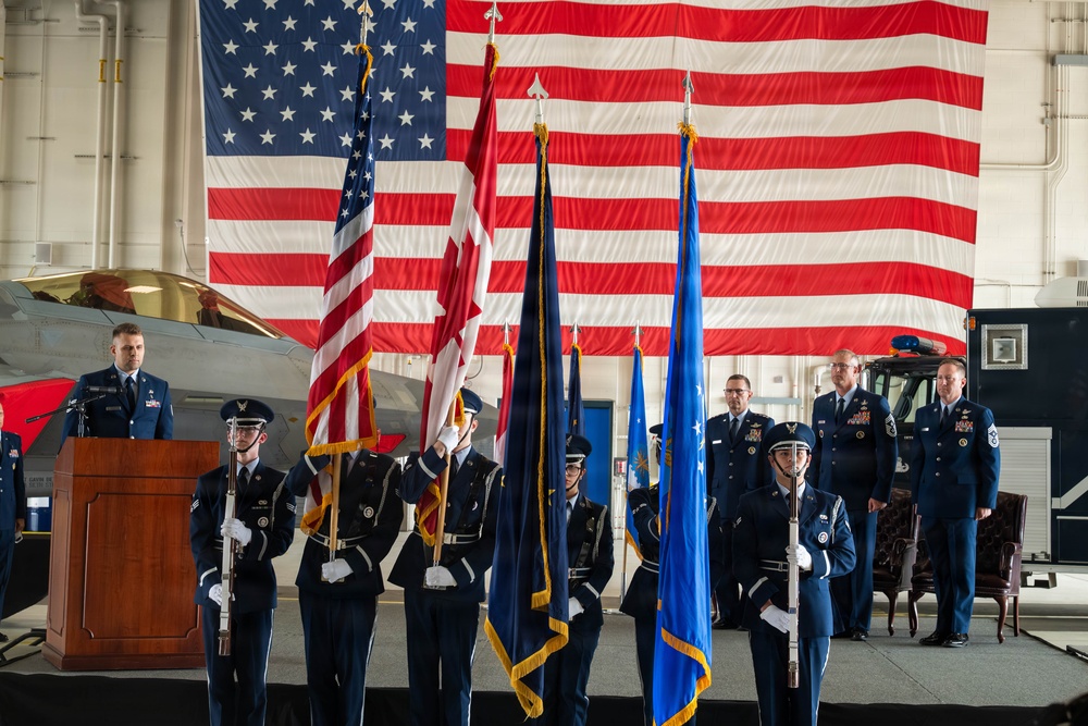 A formal U.S. Air Force military ceremony is taking place inside an aircraft hangar, featuring a color guard presenting flags before a large American flag backdrop. Personnel in dress blue uniforms are positioned at a podium and in formation, with a military aircraft visible to the left. The setting and attire suggest a change of command, activation, or similar official military ceremony.
