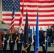 A formal U.S. Air Force military ceremony is taking place inside an aircraft hangar, featuring a color guard presenting flags before a large American flag backdrop. Personnel in dress blue uniforms are positioned at a podium and in formation, with a military aircraft visible to the left. The setting and attire suggest a change of command, activation, or similar official military ceremony.