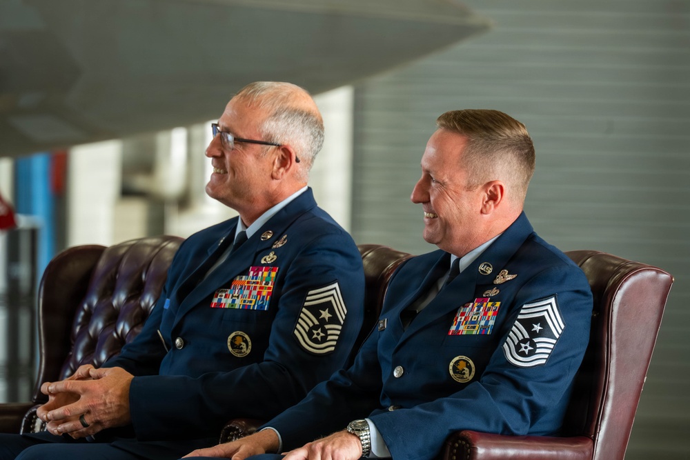 Two senior U.S. Air Force enlisted personnel in dress blue uniforms are seated in leather chairs, both smiling and appearing to enjoy a formal ceremony or event. Both individuals display extensive medal ribbons and Chief Master Sergeant rank insignia on their sleeves. The setting appears to be an indoor military facility, possibly an aircraft hangar or ceremonial hall.