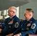 Two senior U.S. Air Force enlisted personnel in dress blue uniforms are seated in leather chairs, both smiling and appearing to enjoy a formal ceremony or event. Both individuals display extensive medal ribbons and Chief Master Sergeant rank insignia on their sleeves. The setting appears to be an indoor military facility, possibly an aircraft hangar or ceremonial hall.