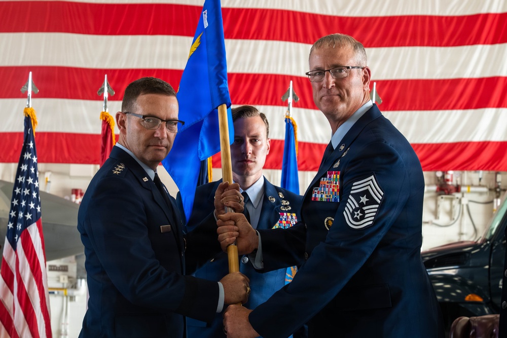 A military ceremony taking place inside an aircraft hangar decorated with a large American flag. Two senior U.S. Air Force officers are exchanging a blue guidon/unit flag in what appears to be a formal change of command or assumption of responsibility ceremony. A third, younger airman stands between them holding the flag staff.