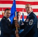 A military ceremony taking place inside an aircraft hangar decorated with a large American flag. Two senior U.S. Air Force officers are exchanging a blue guidon/unit flag in what appears to be a formal change of command or assumption of responsibility ceremony. A third, younger airman stands between them holding the flag staff.