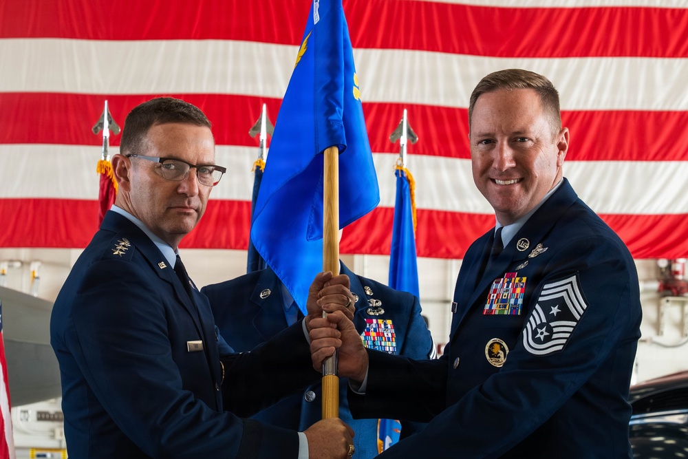 Two U.S. Air Force personnel are participating in a formal guidon/flag transfer ceremony, likely a change of command or assumption of responsibility event. The ceremony is taking place inside a military hangar decorated with a large American flag backdrop. One officer and one senior enlisted member (identifiable by rank insignia) are jointly holding a blue Air Force unit guidon.