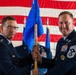Two U.S. Air Force personnel are participating in a formal guidon/flag transfer ceremony, likely a change of command or assumption of responsibility event. The ceremony is taking place inside a military hangar decorated with a large American flag backdrop. One officer and one senior enlisted member (identifiable by rank insignia) are jointly holding a blue Air Force unit guidon.