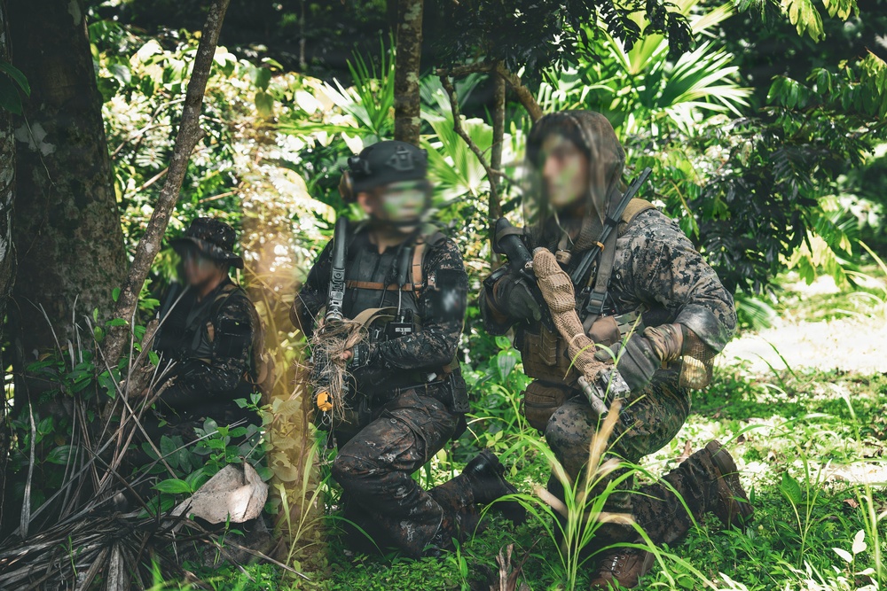 Three armed personnel in military/tactical camouflage gear are positioned in a dense tropical jungle environment. Their faces have been digitally obscured/blurred, suggesting operational security or privacy considerations. The personnel appear to be in a stationary tactical formation among dense vegetation.