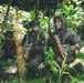 Three armed personnel in military/tactical camouflage gear are positioned in a dense tropical jungle environment. Their faces have been digitally obscured/blurred, suggesting operational security or privacy considerations. The personnel appear to be in a stationary tactical formation among dense vegetation.