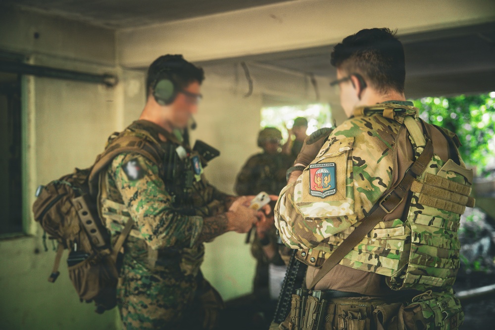 Two military personnel in camouflage uniforms are engaged in a briefing or equipment check inside a covered structure. The soldier facing away displays a patch reading 'AGRUPACION' with what appears to be a parachute insignia, suggesting a special operations or airborne unit. Additional personnel are visible in the background in a similar operational setting.