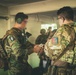 Two military personnel in camouflage uniforms are engaged in a briefing or equipment check inside a covered structure. The soldier facing away displays a patch reading 'AGRUPACION' with what appears to be a parachute insignia, suggesting a special operations or airborne unit. Additional personnel are visible in the background in a similar operational setting.