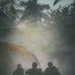 The image shows a group of heavily armed and equipped tactical/military personnel silhouetted against a smoke-filled tropical environment with palm trees and dense vegetation. The figures are viewed from behind, carrying weapons and wearing full tactical gear including helmets. The scene has a dramatic, atmospheric quality due to the heavy smoke or fog diffusing the available light.