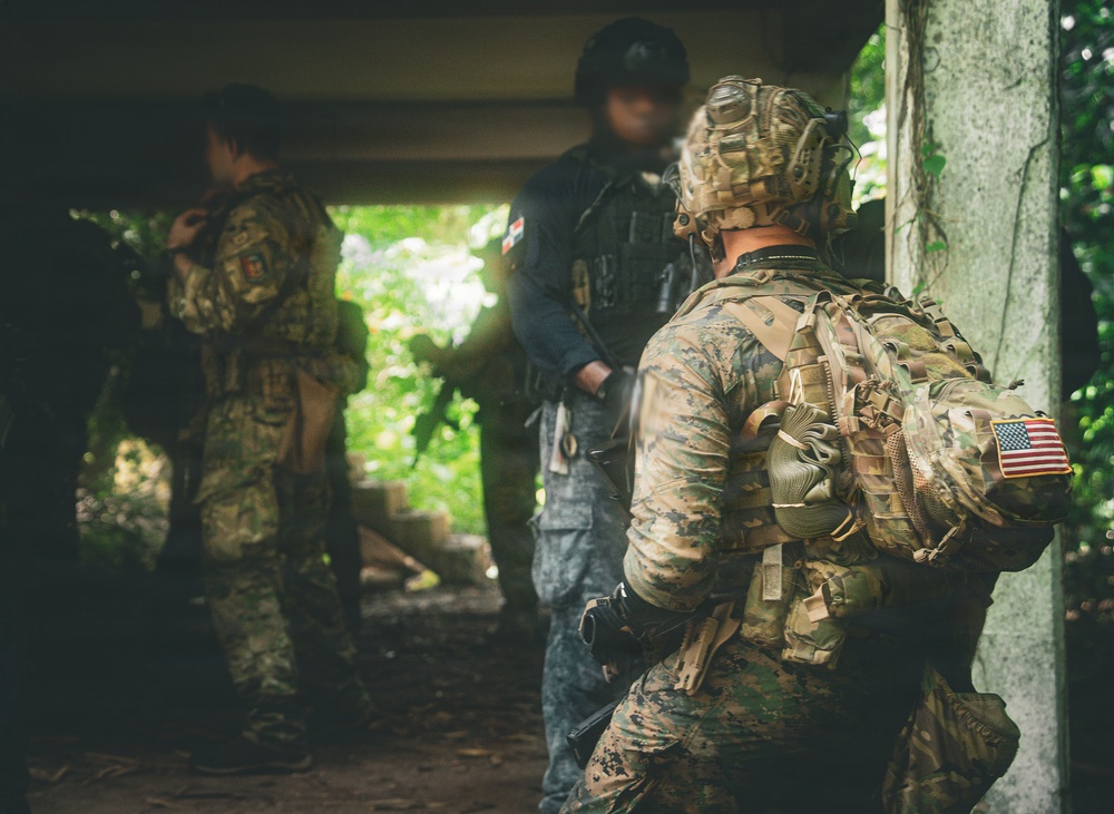 The image shows multiple armed military or law enforcement personnel in a tactical environment, positioned near a concrete structure with dense vegetation visible in the background. The foreground figure wears US Marine Corps MARPAT camouflage with a US flag patch on the backpack and a tactical helmet with night vision mount. The personnel appear to be conducting a coordinated tactical operation or training exercise.