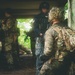 The image shows multiple armed military or law enforcement personnel in a tactical environment, positioned near a concrete structure with dense vegetation visible in the background. The foreground figure wears US Marine Corps MARPAT camouflage with a US flag patch on the backpack and a tactical helmet with night vision mount. The personnel appear to be conducting a coordinated tactical operation or training exercise.