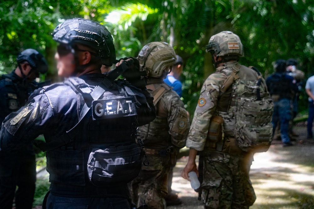 The image shows a group of tactical law enforcement or military personnel gathered in a wooded/jungle environment. At least one officer is clearly identified by the 'G.E.A.T.' patch on their black tactical vest, while others wear multicam/camouflage uniforms with helmets and tactical gear. The personnel appear to be conducting an operation or briefing in an outdoor setting with dense tropical vegetation.