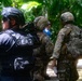 The image shows a group of tactical law enforcement or military personnel gathered in a wooded/jungle environment. At least one officer is clearly identified by the 'G.E.A.T.' patch on their black tactical vest, while others wear multicam/camouflage uniforms with helmets and tactical gear. The personnel appear to be conducting an operation or briefing in an outdoor setting with dense tropical vegetation.