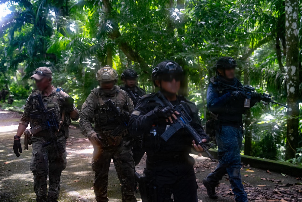 A group of armed military or law enforcement personnel are moving along a paved path through a dense tropical jungle environment. The individuals are wearing mixed uniforms including multicam/camouflage and all-black tactical gear, carrying assault rifles and equipped with body armor and tactical vests. The scene appears to be a joint training exercise or patrol operation in a jungle setting.