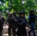 A group of armed military or law enforcement personnel are moving along a paved path through a dense tropical jungle environment. The individuals are wearing mixed uniforms including multicam/camouflage and all-black tactical gear, carrying assault rifles and equipped with body armor and tactical vests. The scene appears to be a joint training exercise or patrol operation in a jungle setting.