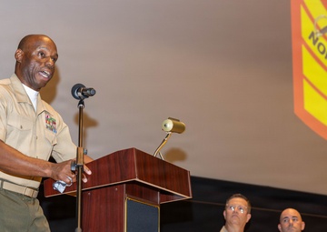 A senior U.S. Marine Corps Staff Noncommissioned Officer is delivering a speech at a podium during an event at the Staff Noncommissioned Officer Academy. He is wearing a tan service uniform with visible rank insignia and service ribbons. Two other Marines in similar uniforms are seated in the background on a stage.