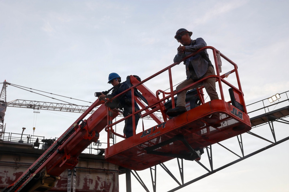 Two workers are elevated on a red JLG boom lift at a construction site. One worker wears a blue hard hat and holds equipment, while the other wears a wide-brimmed hat and appears to be communicating or observing. A large crane and structural steel framework are visible in the background.