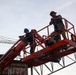 Two workers are elevated on a red JLG boom lift at a construction site. One worker wears a blue hard hat and holds equipment, while the other wears a wide-brimmed hat and appears to be communicating or observing. A large crane and structural steel framework are visible in the background.