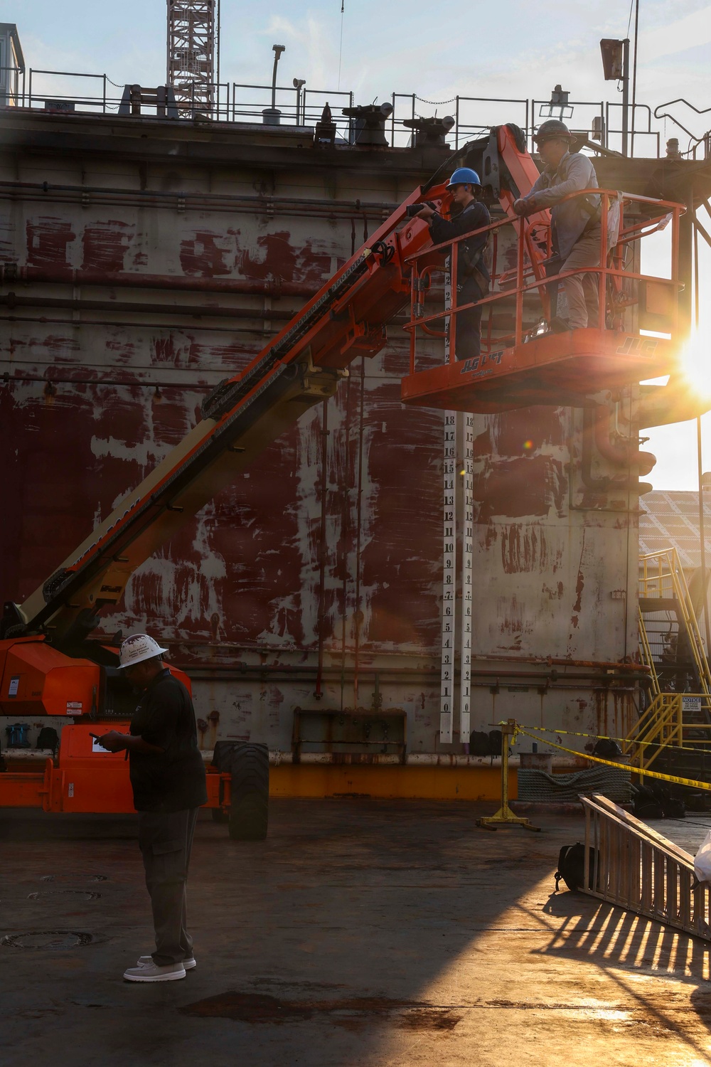 Workers are operating a JLG aerial lift platform alongside a large vessel hull in what appears to be a dry dock or shipyard facility. The hull shows significant weathering with peeling red anti-fouling paint and white primer exposed beneath. A draft measurement scale is visible on the hull, and a supervisor on the ground appears to be reviewing documents or a device.