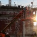 Workers are operating a JLG aerial lift platform alongside a large vessel hull in what appears to be a dry dock or shipyard facility. The hull shows significant weathering with peeling red anti-fouling paint and white primer exposed beneath. A draft measurement scale is visible on the hull, and a supervisor on the ground appears to be reviewing documents or a device.