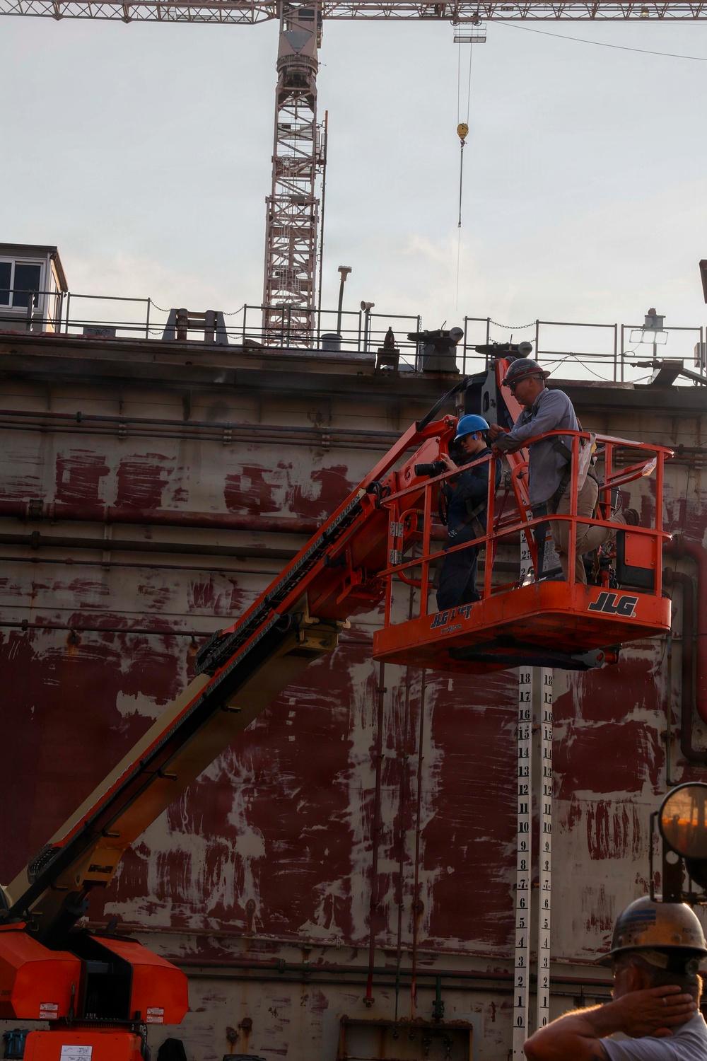 Two workers are positioned in an orange JLG boom lift basket elevated alongside a large vessel or structure with red anti-fouling paint and visible draft markings. A tower crane is visible in the background against an overcast sky. The scene appears to be a shipyard or dry dock facility.
