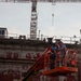 Two workers are positioned in an orange JLG boom lift basket elevated alongside a large vessel or structure with red anti-fouling paint and visible draft markings. A tower crane is visible in the background against an overcast sky. The scene appears to be a shipyard or dry dock facility.