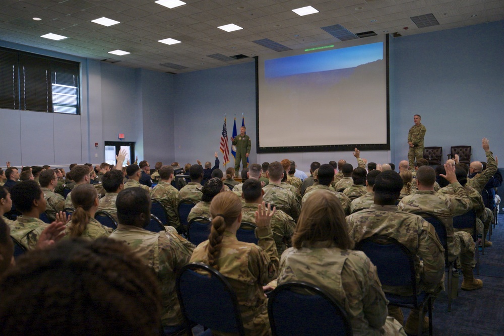A military briefing or all-hands meeting is taking place in a conference room, with a large audience of uniformed personnel seated in rows of chairs. Many audience members have their hands raised, suggesting an interactive Q&amp;A session or polling activity. Two military personnel are standing at the front of the room near a projection screen and American and Air Force flags.