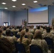 A military briefing or all-hands meeting is taking place in a conference room, with a large audience of uniformed personnel seated in rows of chairs. Many audience members have their hands raised, suggesting an interactive Q&amp;A session or polling activity. Two military personnel are standing at the front of the room near a projection screen and American and Air Force flags.