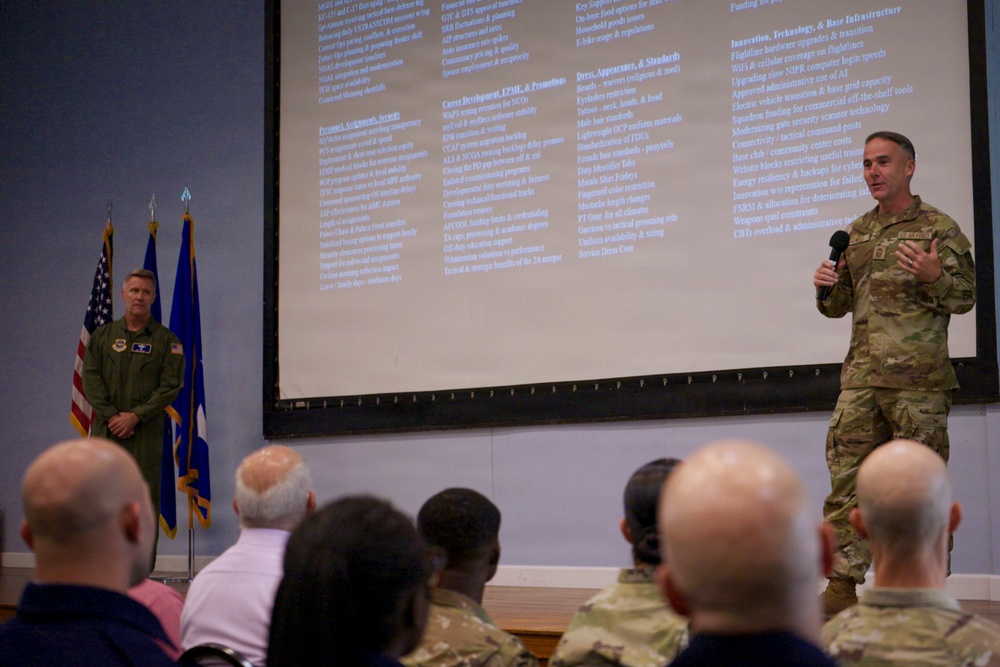A military briefing or town hall meeting is taking place in a conference room setting. One officer in camouflage uniform is speaking into a microphone while another in a green flight suit stands nearby, both positioned in front of a large projection screen displaying multiple categories of topics. An audience of military personnel and civilians is seated facing the presenters.