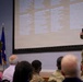 A military briefing or town hall meeting is taking place in a conference room setting. One officer in camouflage uniform is speaking into a microphone while another in a green flight suit stands nearby, both positioned in front of a large projection screen displaying multiple categories of topics. An audience of military personnel and civilians is seated facing the presenters.