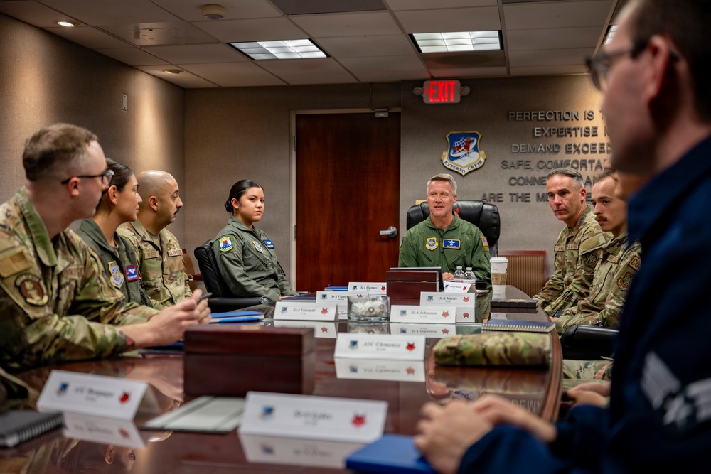 A military briefing or meeting is taking place in a formal conference room, with personnel in various U.S. Air Force uniforms seated around a large reflective table. A senior officer in a green flight suit appears to be leading the discussion from the head of the table. Name placards are visible in front of attendees, and a unit crest with a partially visible motivational quote is displayed on the wall behind the leader.